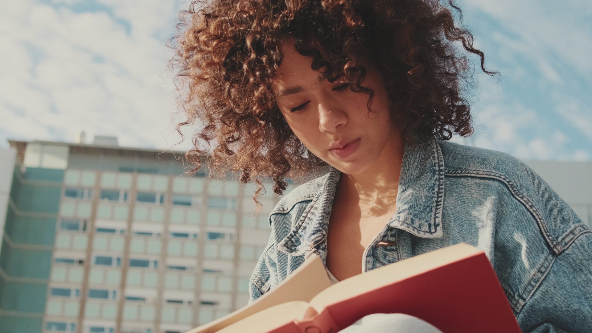 A student reading a book outside a university building on a sunny day.