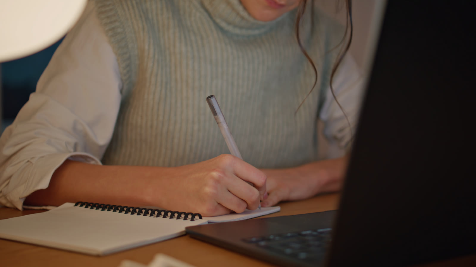 A student writing in a notebook beside a laptop late in the evening.