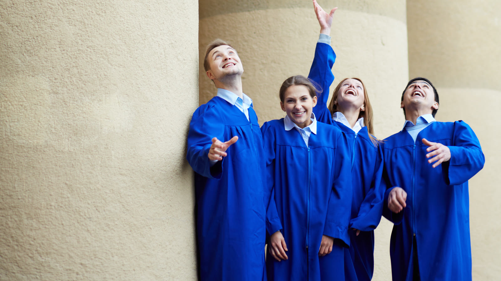 Graduates in blue gowns looking up as they throw their caps into the air.