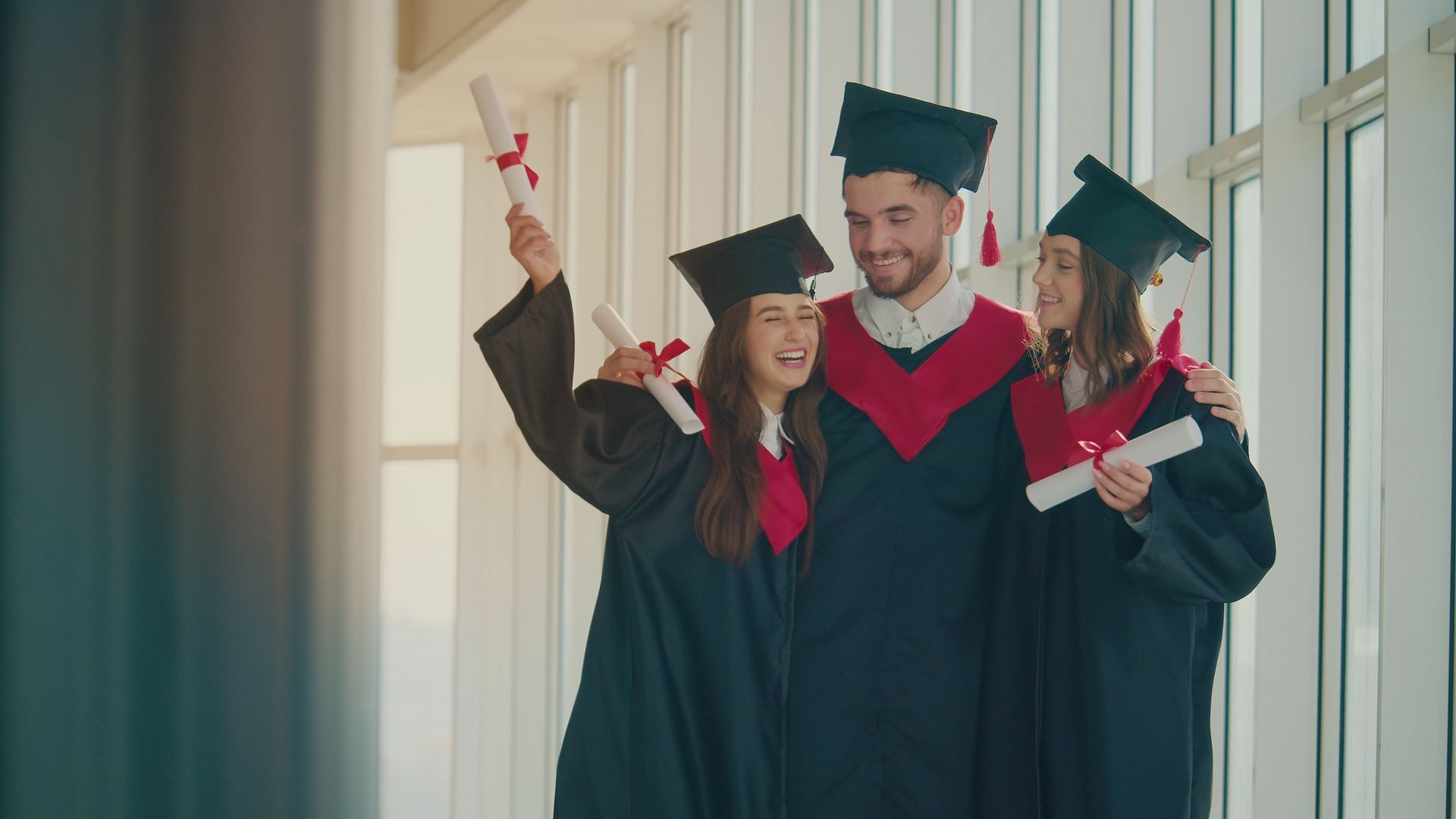 Three happy graduates in caps and gowns hugging with their diplomas