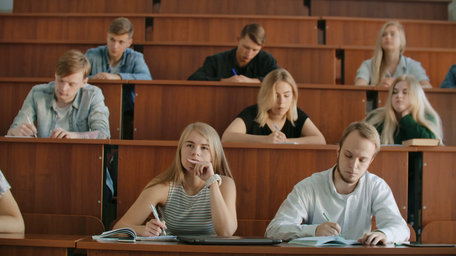 Students in a lecture hall writing in their notebooks while an instructor teaches.